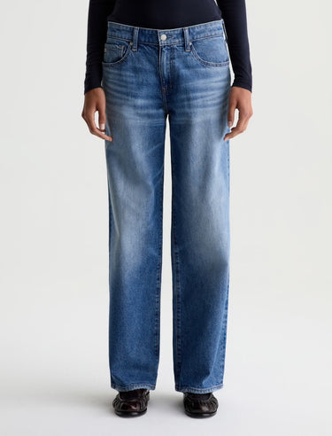A person wearing a pair of medium-wash blue denim jeans with a straight-leg fit, paired with a black long-sleeve top and dark brown shoes, shown against a plain light gray studio background.