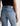 Close-up back view of a person wearing light-wash blue jeans with a black top, showing the back pockets and a tan leather brand patch at the waistband against a plain light background.