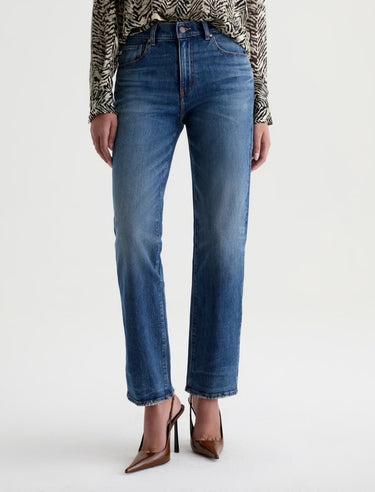 A woman wearing mid-rise medium blue wash straight leg denim with a distressed hem, fading, and gold stitching on a white background.