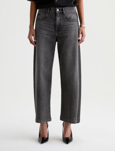 A woman wearing dimensional ashy black wash jeans, with contrasting paneled detailing and a black top with black heels on a white background.
