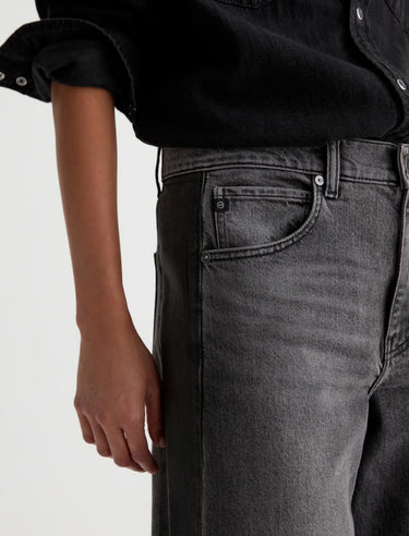 A woman wearing dimensional ashy black wash jeans, with contrasting paneled detailing and a black top on a white background.