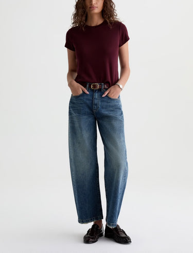 A woman wearing high rise barrel jeans in medium wash with a short sleeve maroon tee and black loafers on a white background.