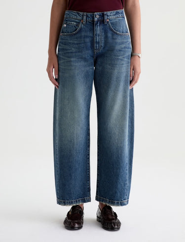 A woman wearing high rise barrel jeans in medium wash with a short sleeve maroon tee and black loafers on a white background.
