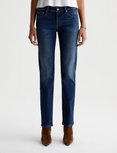 A person wearing dark blue straight-leg denim jeans with a faded wash, paired with a white top and brown suede ankle boots, shown against a plain light gray studio background.