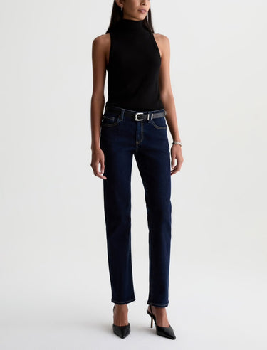 A woman wearing low rise straight dark blue jeans and black heels on a white background.