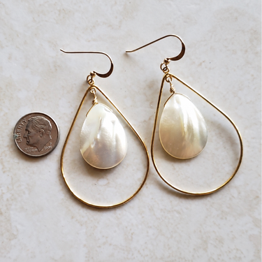 A pair of gold teardrop hoop earrings with white mother-of-pearl teardrop pendants, displayed on a light beige textured surface next to a US dime for scale.