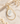 A gold teardrop earring with a white mother-of-pearl teardrop stone, styled on a light beige marble surface with dried flowers.