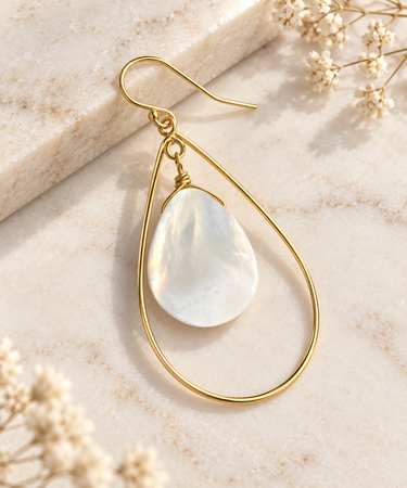 A gold teardrop earring with a white mother-of-pearl teardrop stone, styled on a light beige marble surface with dried flowers.