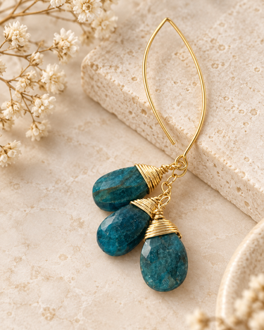 A pair of gold hoop earrings with faceted teal-blue stone drops, styled on a textured beige stone slab with dried white flowers.
