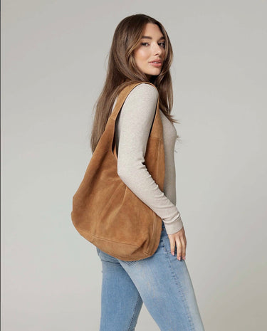 A woman wearing a light beige long-sleeve top and light blue jeans carries a large brown suede shoulder bag against a plain light gray studio background.