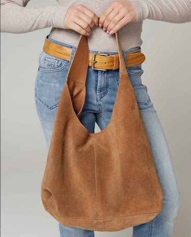 A woman holding a slouchy brown suede hobo shoulder bag with a single strap on a neutral background