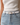 A close-up of a model wearing a slim white leather belt with a gold-tone rectangular buckle, styled with light-wash blue jeans and a white ribbed top.