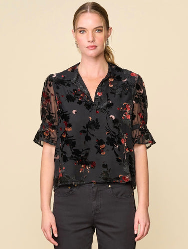 A woman wearing a velvet floral mesh blouse and black jeans on a neutral background.