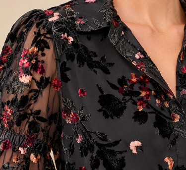 A close up of woman wearing a velvet floral mesh blouse and black jeans on a neutral background.