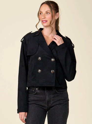 A woman modeling a black double-breasted cropped jacket with silver buttons, paired with dark black jeans, against a solid light beige background.