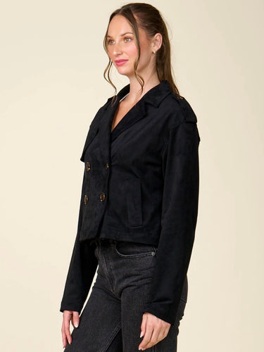 A woman modeling a black double-breasted cropped jacket with silver buttons, paired with dark black jeans, against a solid light beige background.