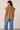 A woman shown from the back wearing a short-sleeve brown top with a gathered neckline and blue jeans, standing against a plain light beige wall.