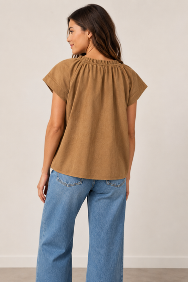 A woman shown from the back wearing a short-sleeve brown top with a gathered neckline and blue jeans, standing against a plain light beige wall.