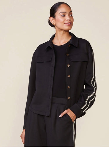 A woman modeling a black cropped button-front jacket with white double stripes down the sleeves, paired with matching black pants, against a plain beige studio background.