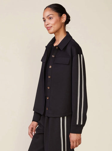 A woman modeling a black cropped button-front jacket with white double stripes down the sleeves, paired with matching black pants, against a plain beige studio background.
