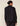 A woman shown from the back wearing a black long-sleeve collared jacket with subtle sheer side panels and black pants, photographed against a plain light beige studio background.