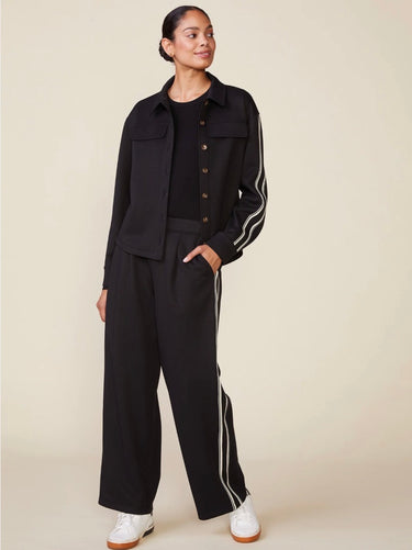 A model wearing a black two-piece tracksuit with white double stripes down the sleeves and legs, featuring a cropped button-front jacket and matching wide-leg pants, paired with white sneakers against a solid beige background.
