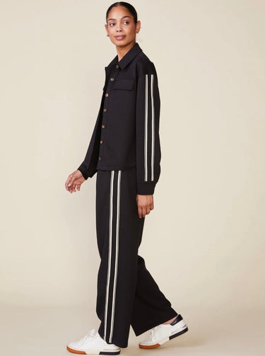 A model wearing a black two-piece tracksuit with double white stripes down the sleeves and legs, paired with white sneakers, against a solid beige background.