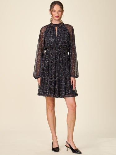 A woman modeling a short black mesh dress with a white polka dot pattern, long sheer sleeves, and a high keyhole neckline, paired with black slingback heels against a solid beige background.