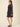 A woman modeling a short black dress with a small white polka dot pattern, featuring long sheer bell sleeves and a keyhole neckline, paired with black slingback heels against a solid beige background.
