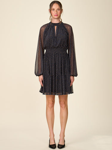A woman modeling a sheer long-sleeve mini dress with a small white polka dot pattern on a black base, featuring a high keyhole neckline and a flared skirt, set against a solid beige studio background.