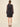 A model viewed from the back wearing a sheer black long-sleeve mini dress with a small white polka dot pattern, a keyhole back, and a tiered skirt, paired with black high heels against a solid beige background.