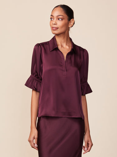 A woman modeling a dark plum satin blouse with short ruffled sleeves and a split V-neckline, paired with a matching satin skirt against a solid beige background.