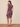 A woman modeling a sleeveless knee-length dress with a floral print, shown from the back. She is holding a small pink clutch and wearing beige high-heeled sandals against a plain beige studio backdrop.