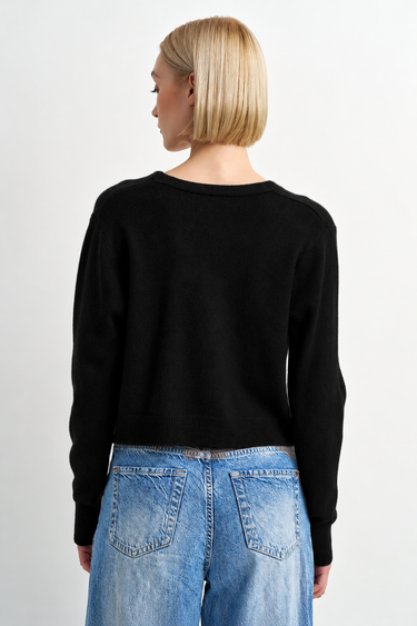 A woman wearing a black cardigan and blue jeans on a white background.