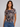 A woman modeling a short-sleeve crewneck t-shirt with a muted geometric block pattern in gray, brown, and rust tones, paired with blue jeans against a solid light gray background.