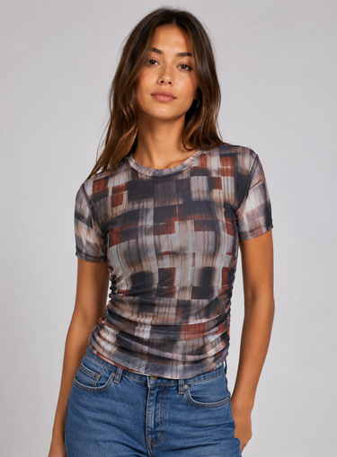 A woman modeling a short-sleeve crewneck t-shirt with a muted geometric block pattern in gray, brown, and rust tones, paired with blue jeans against a solid light gray background.