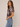 A woman modeling a short-sleeve plaid top in muted brown, gray, and white tones, paired with light blue jeans, against a plain beige studio background.