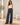 A woman stands in a bright, modern room wearing high-waisted, wide-leg navy pinstripe trousers with a gold-tone belt, paired with a white tank top and black loafers.