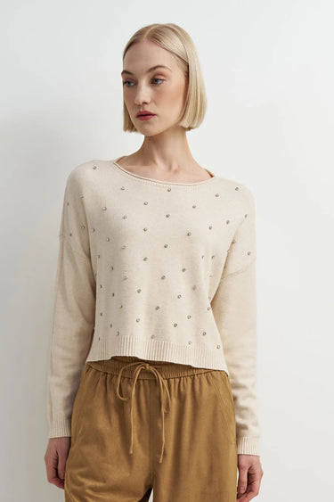 A woman wearing an embellished cream knit sweater and mustard yellow pants on a white background.