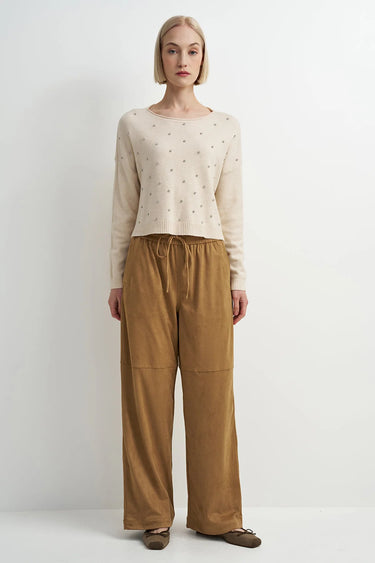 A woman wearing an embellished cream knit sweater and mustard yellow pants on a white background.