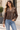 A woman wearing a brown faux-leather jacket with a tan sherpa collar and cuffs, paired with light blue jeans, standing outdoors in front of a beige building.