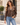 A woman wearing a brown faux-leather jacket with a tan sherpa collar and cuffs, paired with light blue jeans, standing outdoors in front of a beige building.