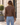 A woman viewed from behind wearing a brown faux-leather jacket with a tan sherpa collar and cuffs, paired with light blue jeans, standing outdoors in front of a beige building.