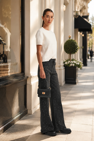 A woman stands on a sunlit city sidewalk wearing a white short-sleeve knit top, dark wide-leg embellished jeans, black shoes, and holding a small black handbag.