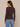 A woman shown from the back wearing a dark brown ribbed long-sleeve sweater and light blue jeans, standing against a plain beige studio background.