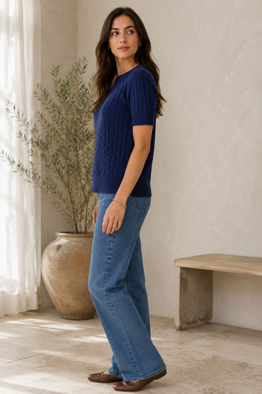 A woman wearing a navy blue short-sleeve cable-knit top and light blue wide-leg jeans, standing in a bright, neutral-toned room with a potted plant and a wooden bench.