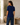 A woman wearing a short-sleeve navy blue ribbed knit top and dark blue jeans, photographed from the back in a bright, modern room with a window, potted plant, and wooden bench.