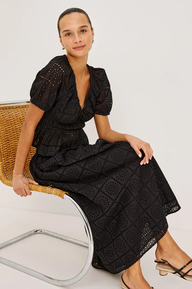 woman wearing black embroidery blouse and skirt seated on a chair on white background