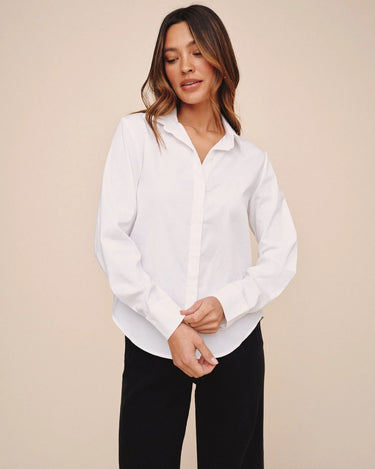 A woman wearing a white long-sleeve button-up shirt and black pants, standing against a solid beige studio background.