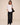 A woman modeling a white long-sleeve button-down shirt and black wide-leg trousers, carrying a black shoulder bag against a solid beige studio background.